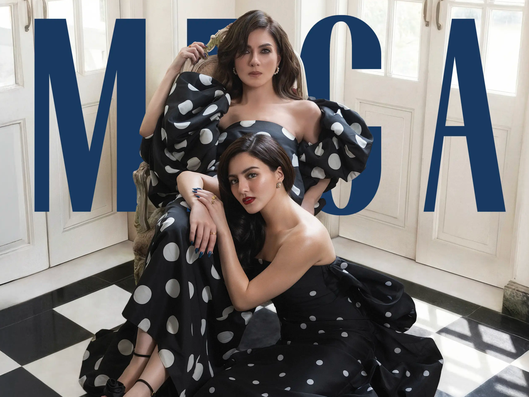Two women in black polka-dot gowns pose closely in a white, tiled room with large blue letters behind them (fashion editorial).